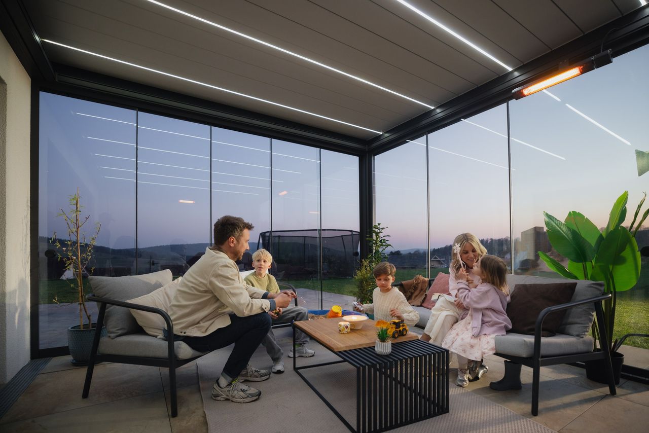 PERGOLUX Lamellendach mit LED in den Lamellen und Glaswänden am Abend, darunter eine Familie