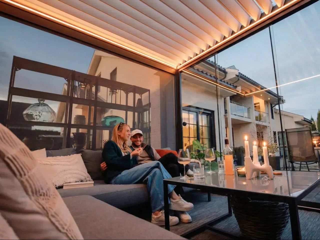 PERGOLUX Lamellendach mit LED-Beleuchtung am Abend, darunter zwei Personen auf einem Sofa
