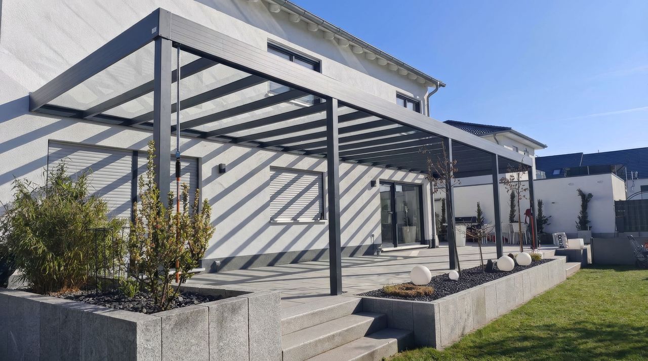 Terrassenüberdachung aus Aluminium in Anthrazit mit Glasdach über einer langen Terrasse an einem weißen Haus